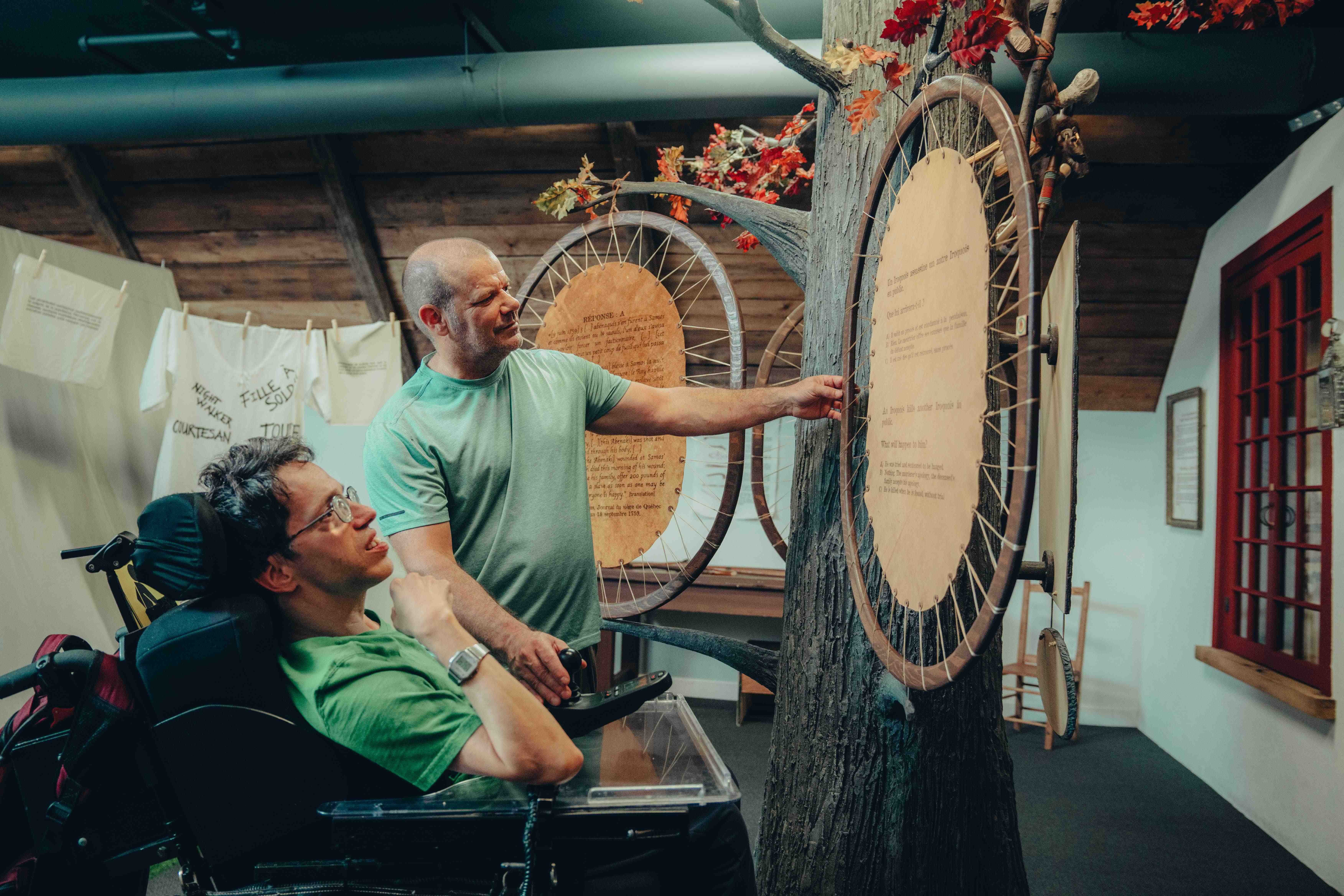 Homme en fauteuil roulant accompagné visitant l'exposition du Musée des plaines d'Abraham. // A man in a wheelchair accompanied by someone visiting the exhibition at the Plains of Abraham Museum.