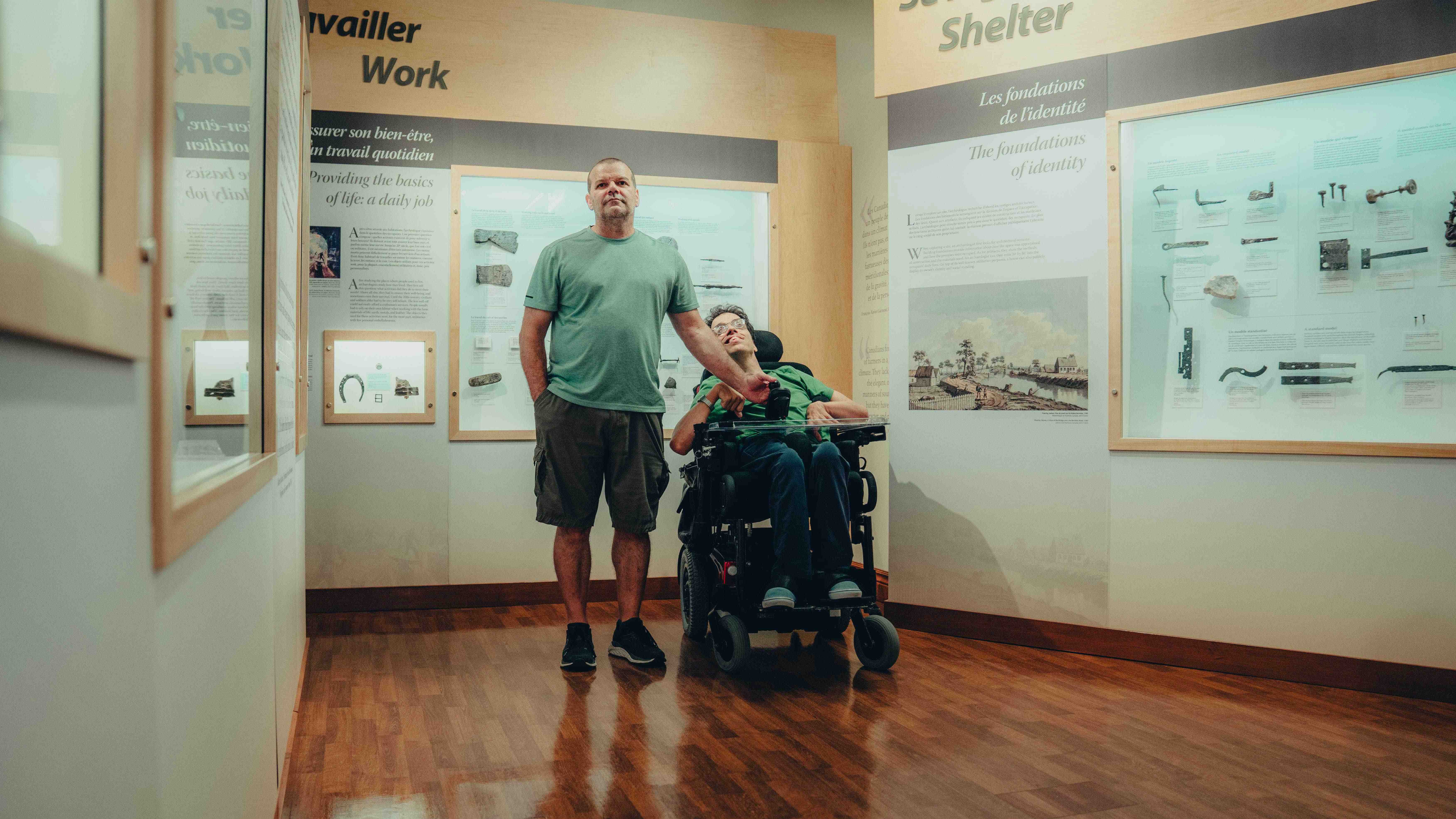 Homme en fauteuil roulant accompagné déambulant dans l'exposition du Musée des plaines d'Abraham. // A man in a wheelchair accompanied by someone strolling through the exhibition at the Plains of Abraham Museum.