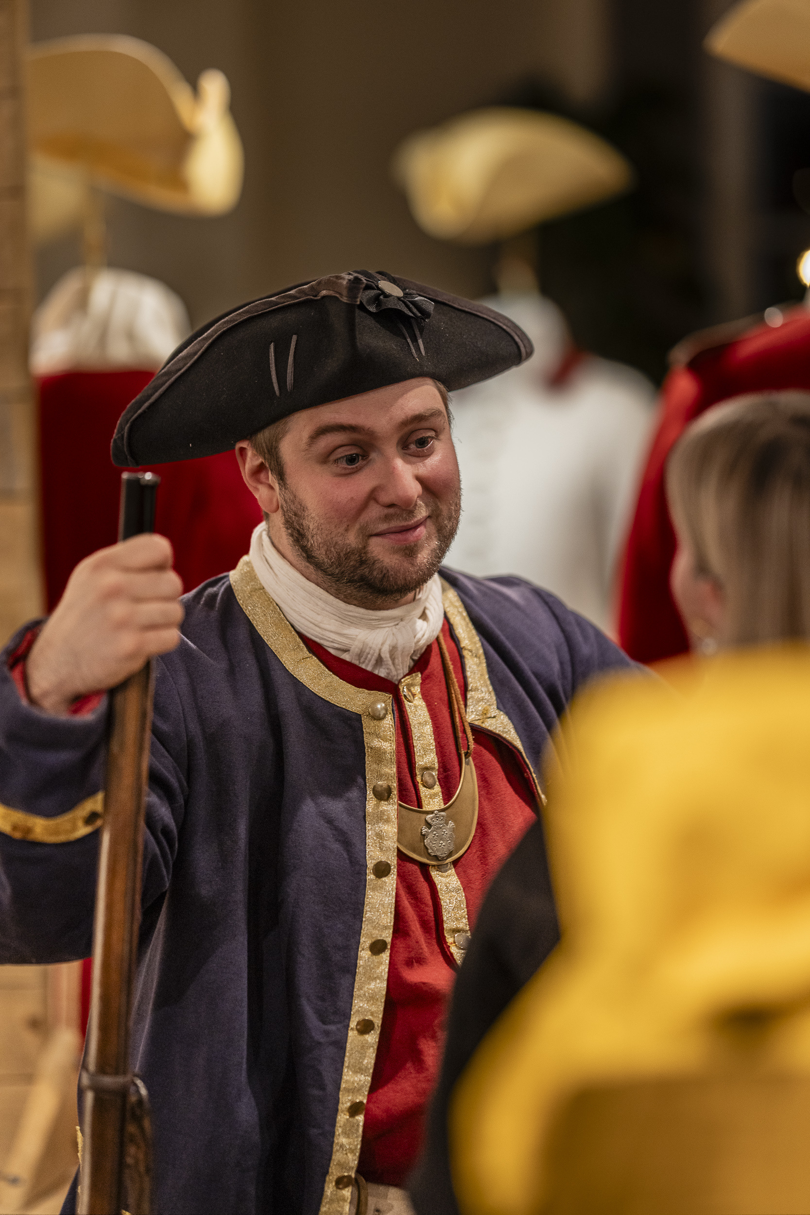 Homme costumé portant un chapeau noir et une veste de couleur bleu, jaune et rouge et tenant un fusil. Man in costume wearing a black hat and a blue, yellow, and red jacket, holding a rifle.