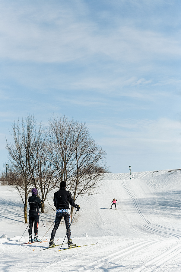 Deux skieurs vêtus en noir posant de dos sur les pistes de fond des plaines d'Abraham, on y voit devant eux un skieur en train de monter la pente. // Two skiers dressed in black posing from behind on the cross-country ski trails of the Plains of Abraham, with a skier in front of them going up the hill.