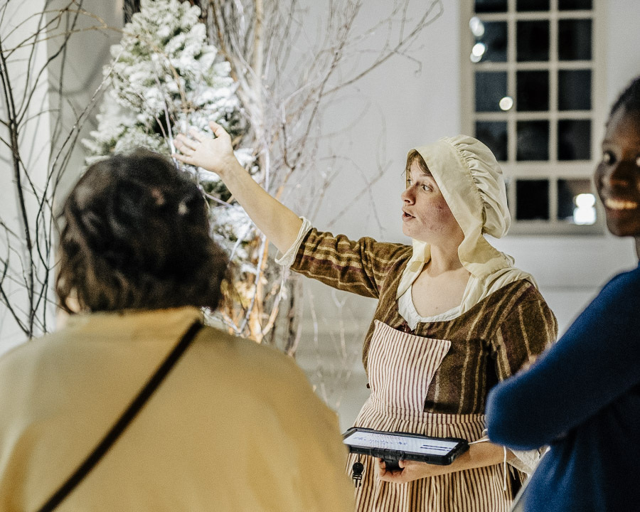 Une femme portant un costume d'époque et tenant une tablette raconte à deux femmes une partie de l'exposition.