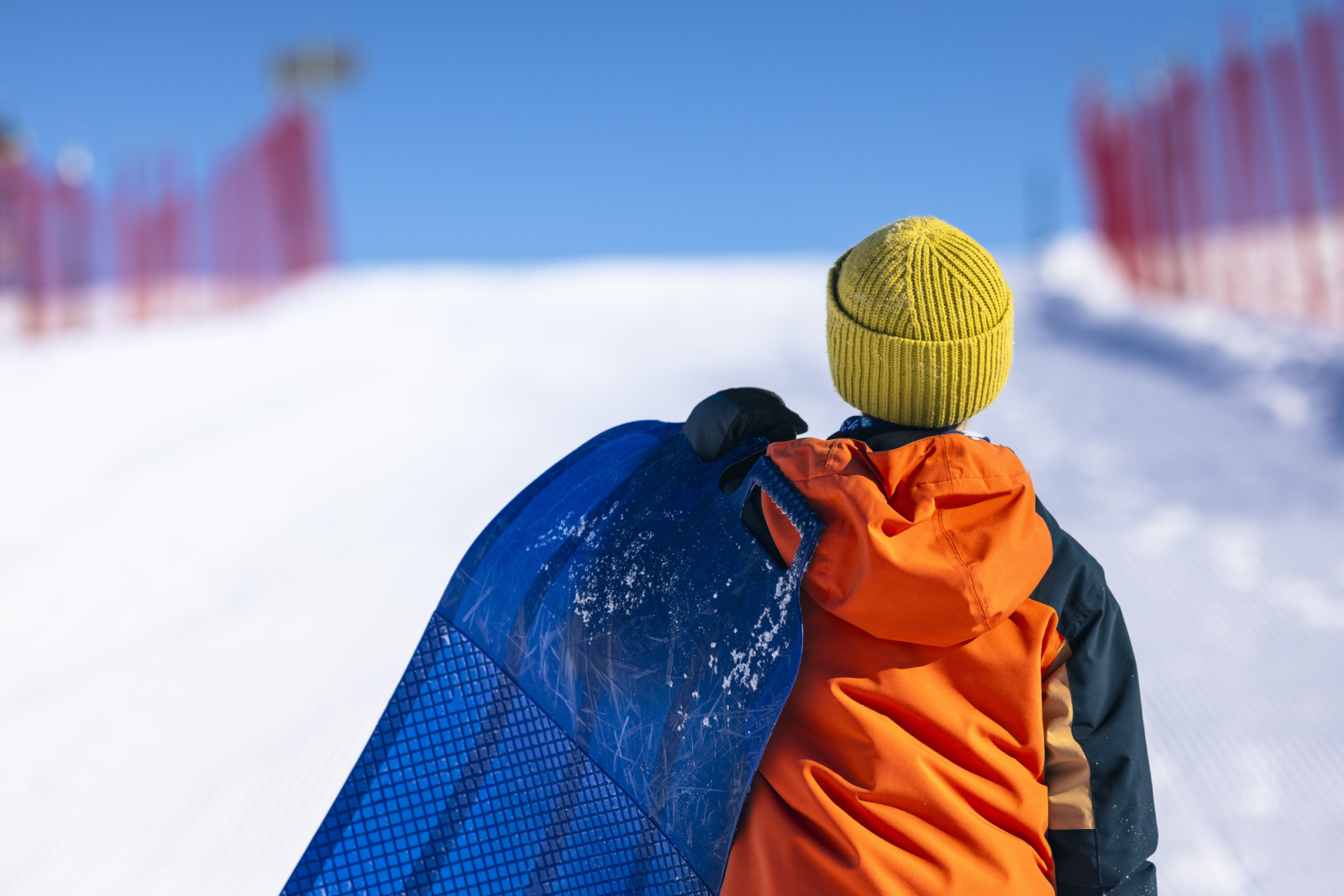 Enfant portant un manteau orange et une tuque jaune, vu de dos, tenant un tapis à neige au sommet d’une glissade sur les plaines d’Abraham. // Child in an orange coat and yellow winter hat, seen from behind, holding a snow carpet at the top of a slide on the Plains of Abraham.