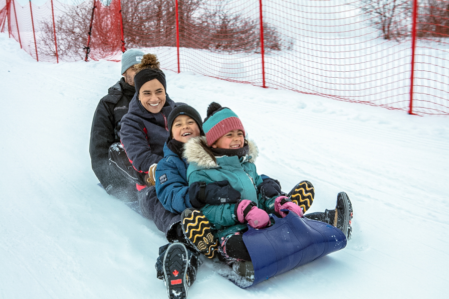 Une famille de 2 adultes et 2 enfants glissant sur un tapis à neige sur les plaines d'Abraham. A family of 2 adults and 2 children sliding on a snow carpet on the Plains of Abraham.