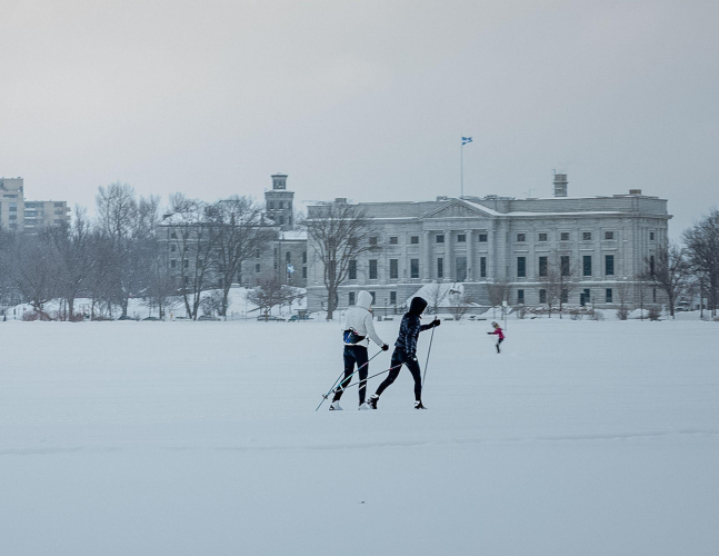 Deux skieurs sur les plaines d'Abraham par une journée enneigée, on aperçoit l'ancien bâtiment du Musée national des beaux-arts du Québec derrière eux. Two skiers on the Plains of Abraham on a snowy day, with the old building of the Musée national des beaux-arts du Québec visible behind them.