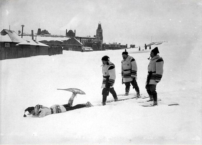 Quatre hommes portant un habit de neige identique chaussant des raquettes sur les plaines d'Abraham, l'un d'entre eux est couché au sol avec une jambe et une raquette dans les airs après être tombé. Four men wearing identical snow outfits and snowshoes on the Plains of Abraham, one of them lying on the ground with one leg and a snowshoe in the air after falling.