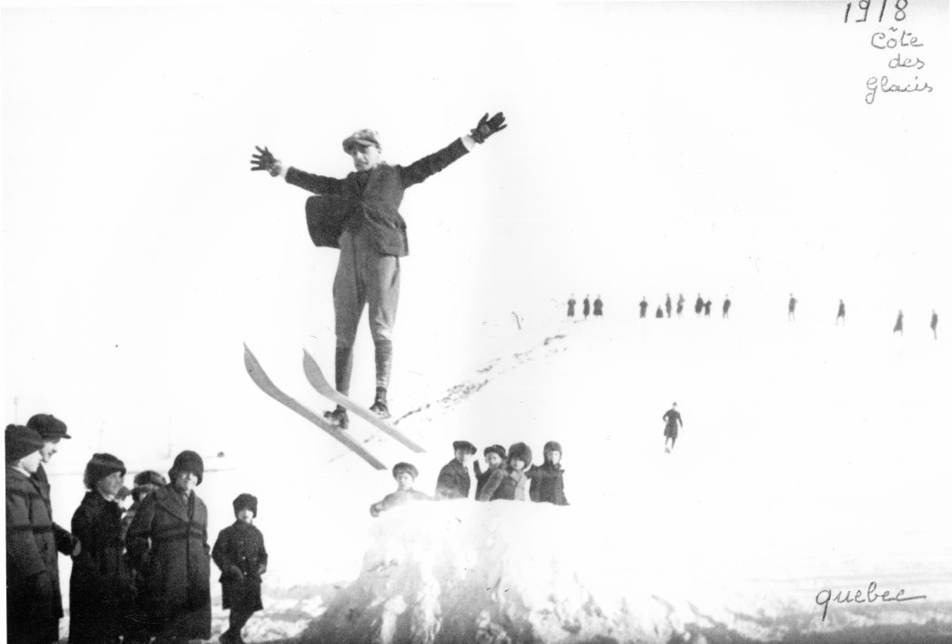 Photo d'archive datant de 1918, on y aperçoit un skieur effectuant un saut les bras dans les airs. Archive photo from 1918 showing a skier mid-jump with arms in the air.