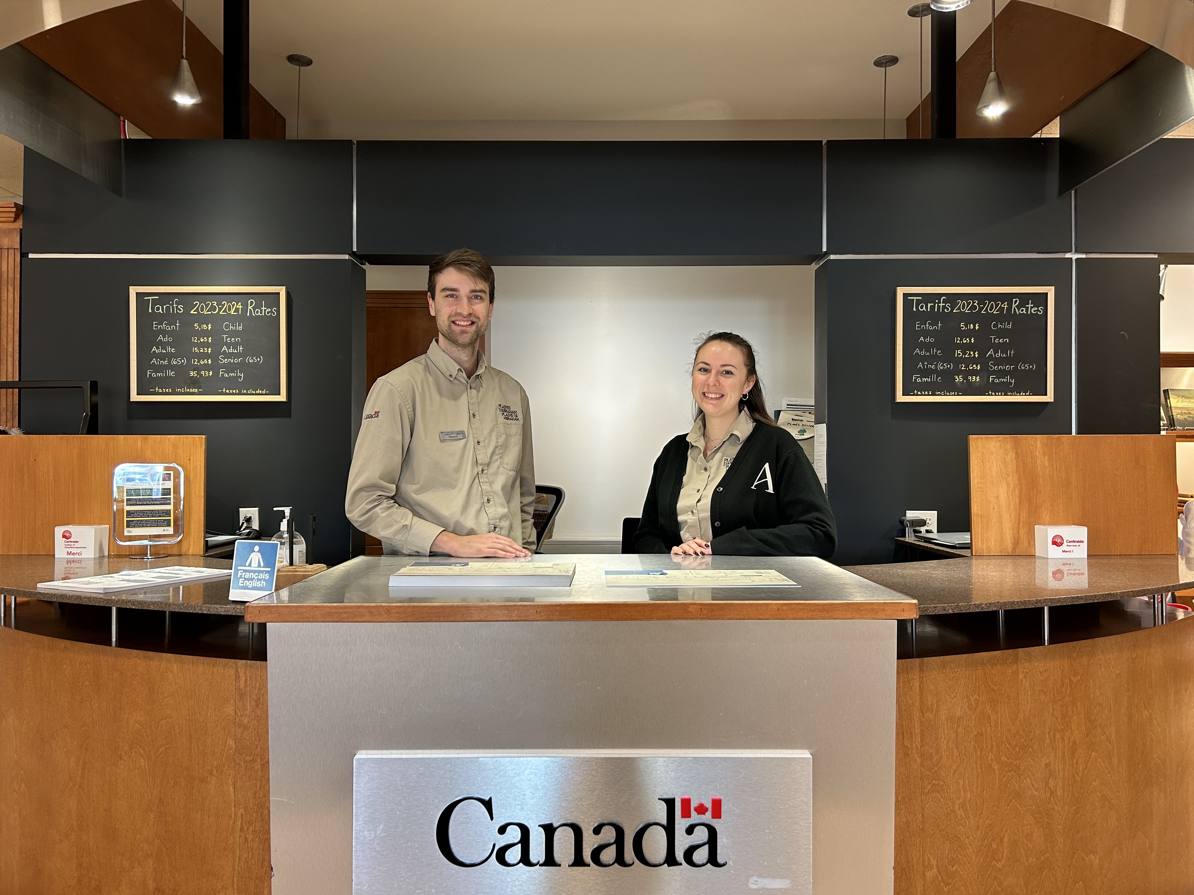 Un homme et une femme derrière le comptoir d'accueil du Musée des plaines d'Abraham. A man and a woman standing behind the reception desk at the Plains of Abraham Museum.