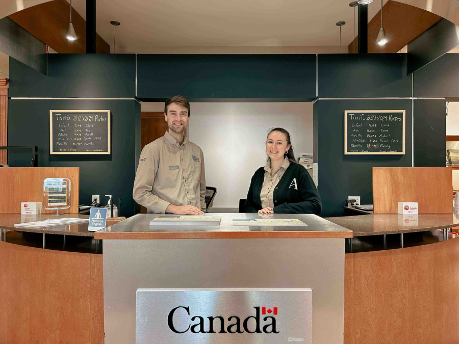 Un homme et une femme derrière le comptoir d'accueil du Musée des plaines d'Abraham. A man and a woman standing behind the reception desk at the Plains of Abraham Museum.