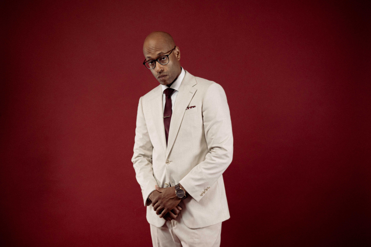 Artiste Webster posant en smoking beige sur fond rouge bourgogne.