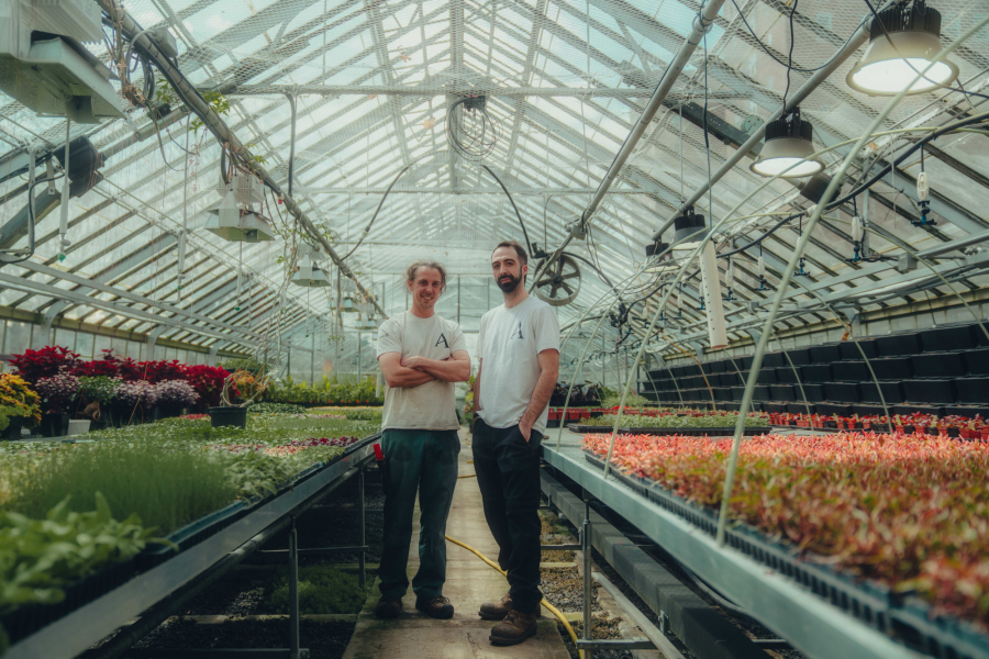 Deux employés de l'équipe horticole des plaines d'Abraham posant dans l'allée centrale de la serre, semis déposés des deux côtés de l'allée. Two employees from the Plains of Abraham horticultural team posing in the central aisle of the greenhouse, with seedlings placed on both sides of the aisle.