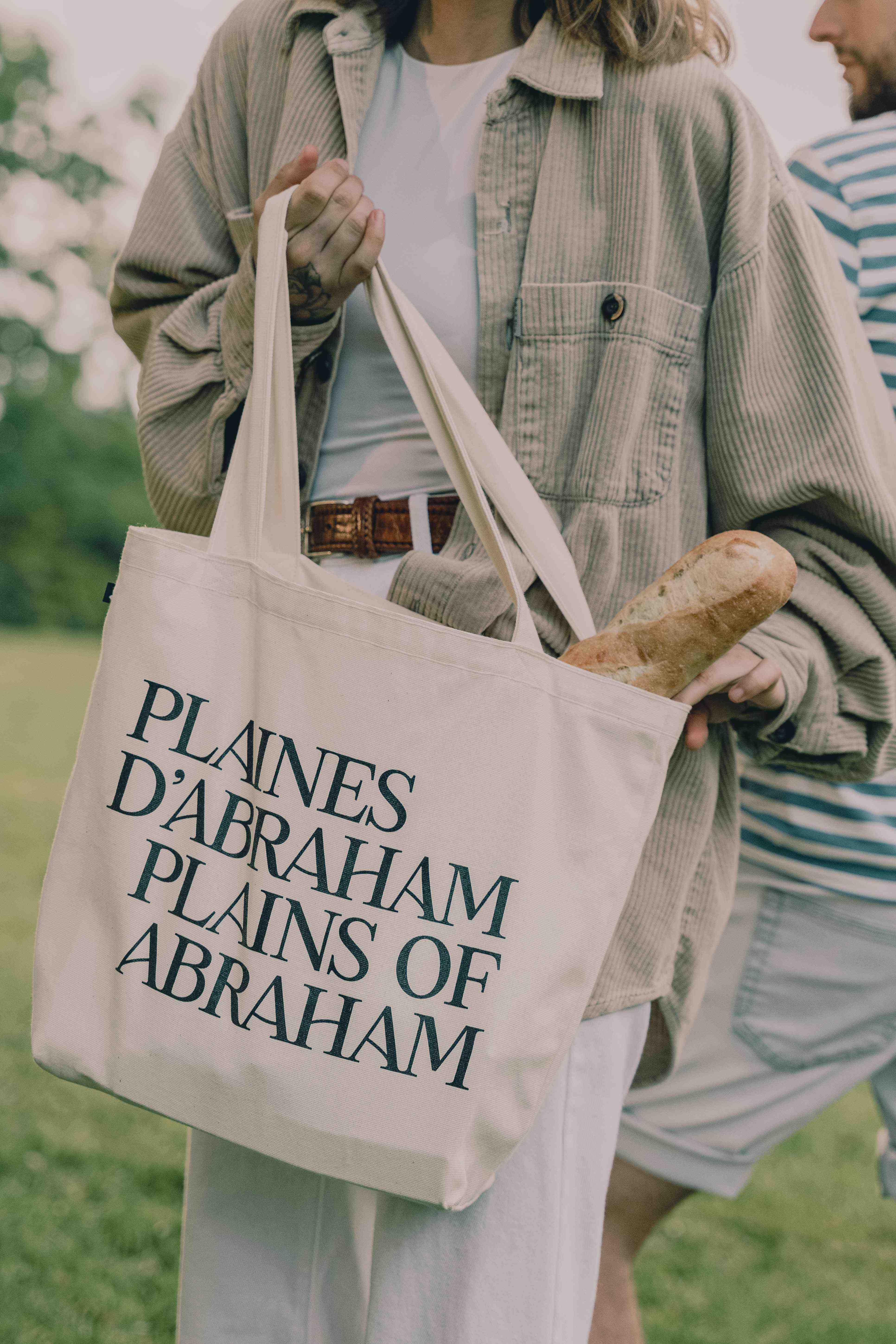 Photo d'une femme avec un sac beige à la main lettré Plaines d'Abraham, le sac contient une miche de pain. On aperçoit aussi son conjoint derrière elle. // Photo of a woman holding a beige bag labeled "Plaines d'Abraham," with a loaf of bread inside. Her partner can also be seen behind her.