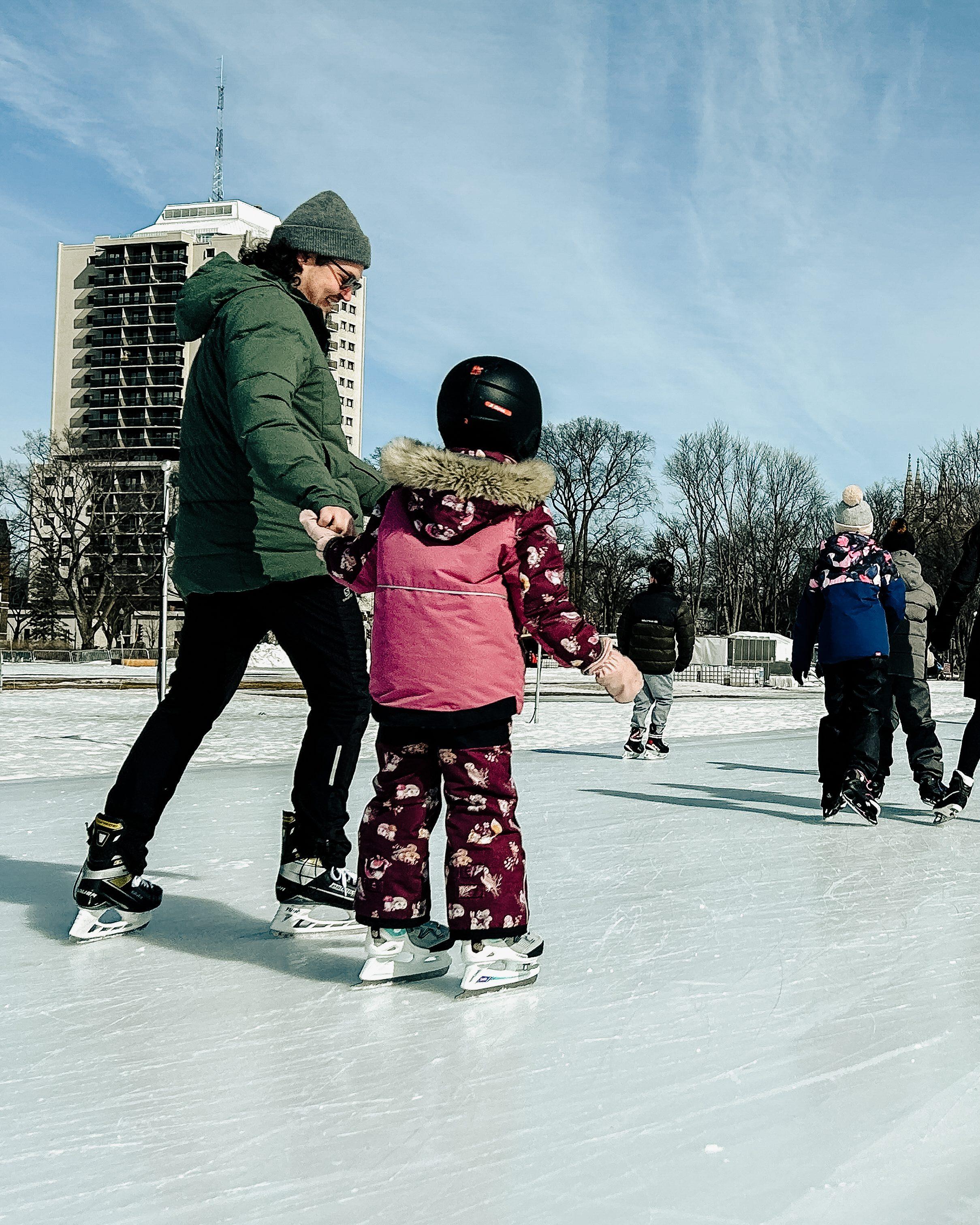 Un père et sa fille patinant sur l'anneau de glace des plaines d'Abraham, le père porte un manteau vert et sa fille un manteau rose.