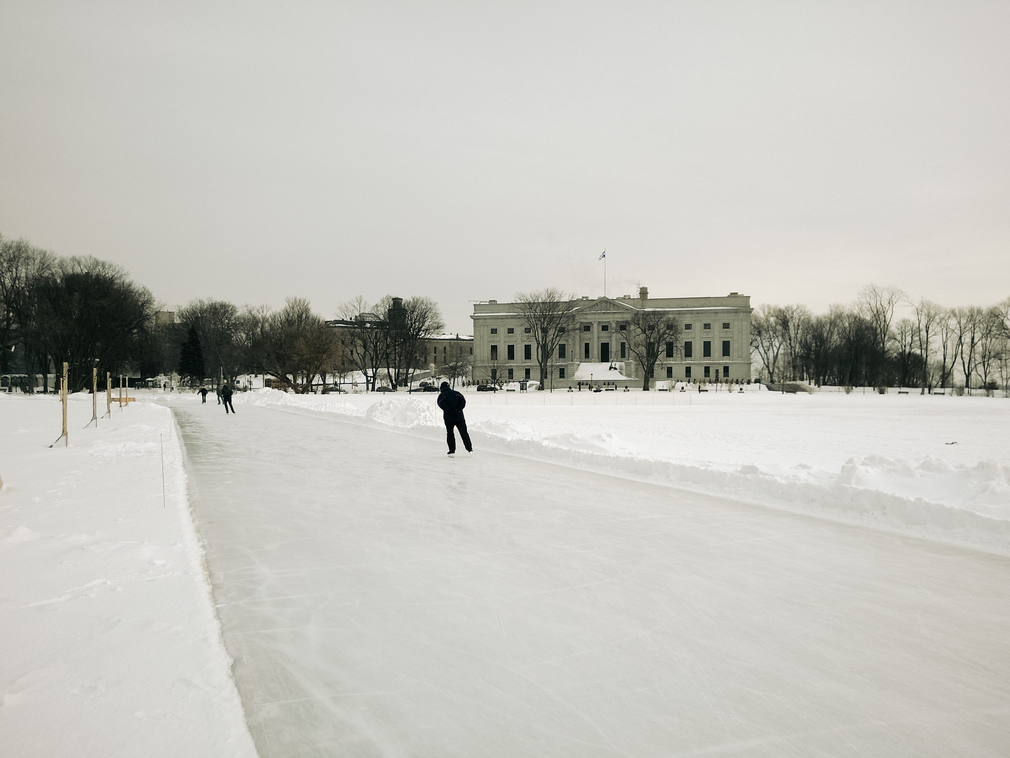 Anneau de glace réfrigéré des plaines d'Abraham, on aperçoit un patineur et un bâtiment du Musée national des beaux-arts du Québec. // Refrigerated skating rink on the Plains of Abraham, featuring a skater and a building of the National Museum of Fine Arts of Quebec.