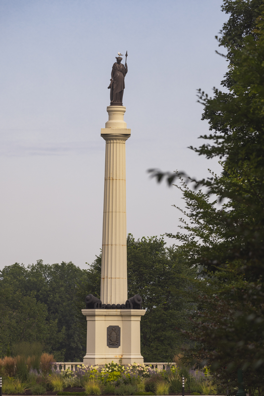 Image du Monument des Braves, situé dans le parc des Braves à Québec, géré par les Plaines d'Abraham. Le monument, d'une hauteur de 22 mètres, commémore la bataille de Sainte-Foy du 28 avril 1760. Il est surmonté d'une statue de Bellone, déesse romaine de la guerre, au sommet d'une colonne, avec des plaques à la base honorant les généraux des armées adverses, Lévis et Murray. Le monument est entouré de verdure luxuriante, soulignant sa signification historique et sa beauté.