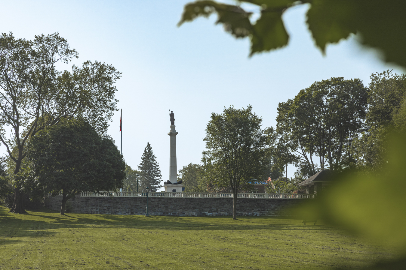 Image of the Monument des Braves located in Parc des Braves, Quebec City, which is owned and managed by the Plains of Abraham. The monument, standing 22 meters high, commemorates the Battle of Sainte-Foy on April 28, 1760. It features a statue of Bellona, the Roman goddess of war, atop a column, with plaques at the base honoring the generals of the opposing armies, Lévis and Murray. The monument is surrounded by lush greenery, highlighting its historical significance and beauty.