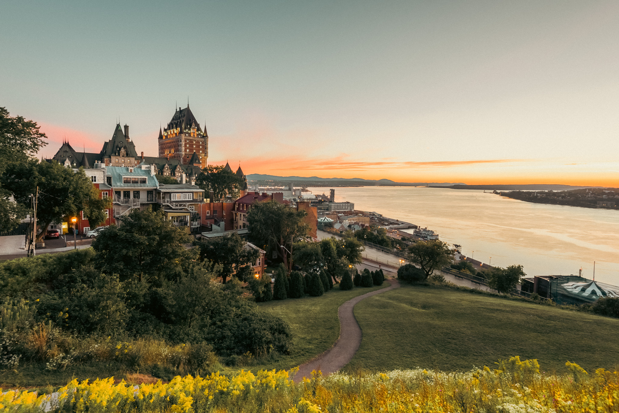 Terrasse Pierre-Dugua-de-Mons en soirée, végétation et maison du Vieux-Québec, vue sur le Château Frontenac de Québec et sur le fleuve Saint-Laurent. Pierre-Dugua-de-Mons Terrace in the evening, with vegetation and a house from Old Quebec, view of the Château Frontenac of Québec City and St-Lawrence River.