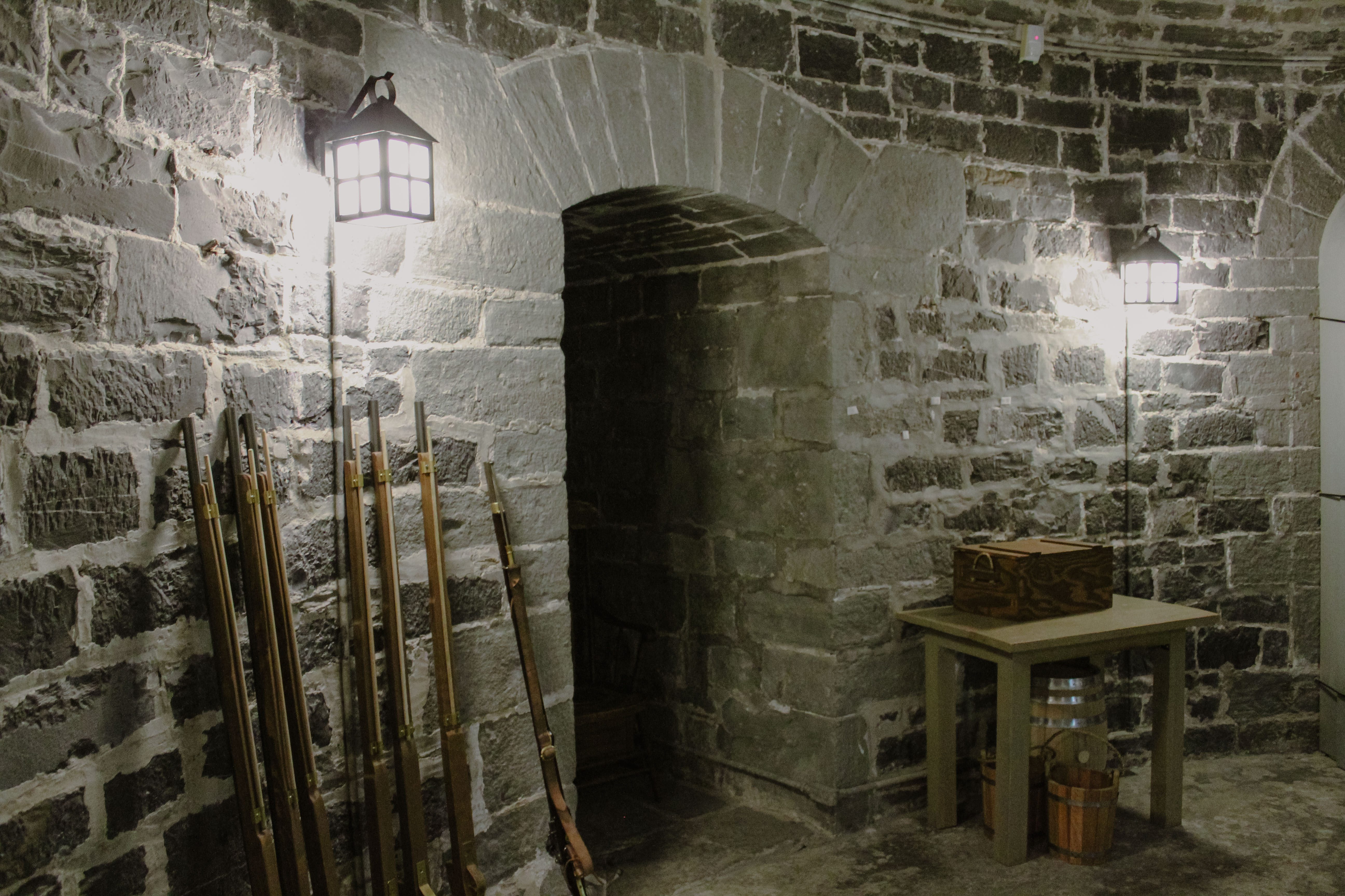 Photo de l'intérieur de la tour Martello 4, mur de pierres, arche et lanternes fixées au mur. Photo of the interior of Martello Tower 4, featuring a stone wall, an arch, and lanterns fixed to the wall.