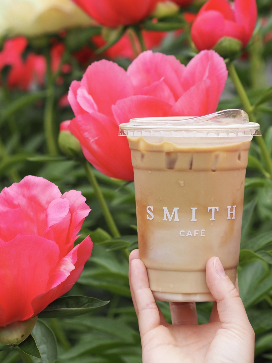 Café glacé de la Maison Smith présenté devant des fleurs de couleur rose. La Maison Smith iced coffee displayed in front of pink flowers.