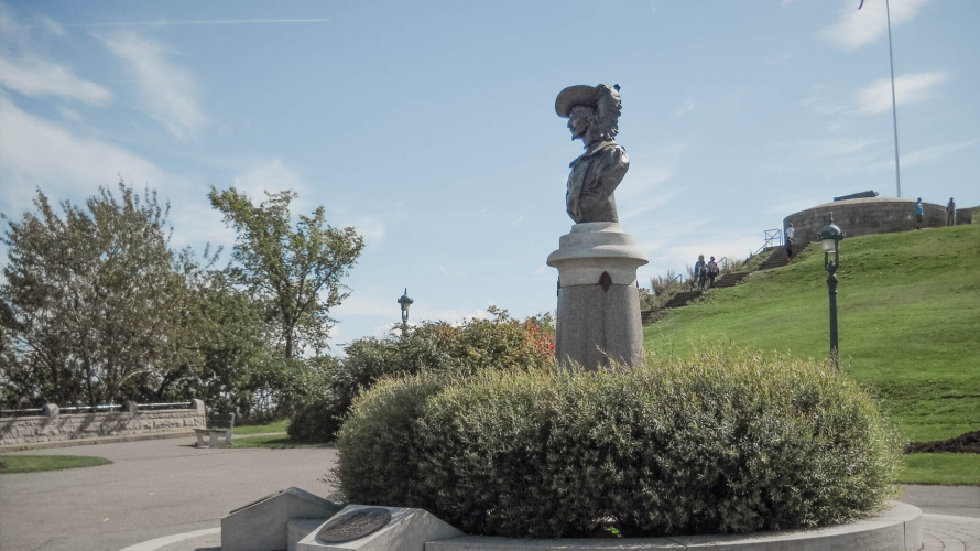 Image du monument Pierre-Dugua-de-Mons situé sur la terrasse Pierre-Dugua-de-Mons, sur le territoire des plaines d'Abraham. Le monument commémore l'importance de la culture française au Canada. Pierre Dugua de Mons est une figure clé de la colonisation de la Nouvelle-France. La statue est placée sur un fond de paysage magnifique des plaines d'Abraham.