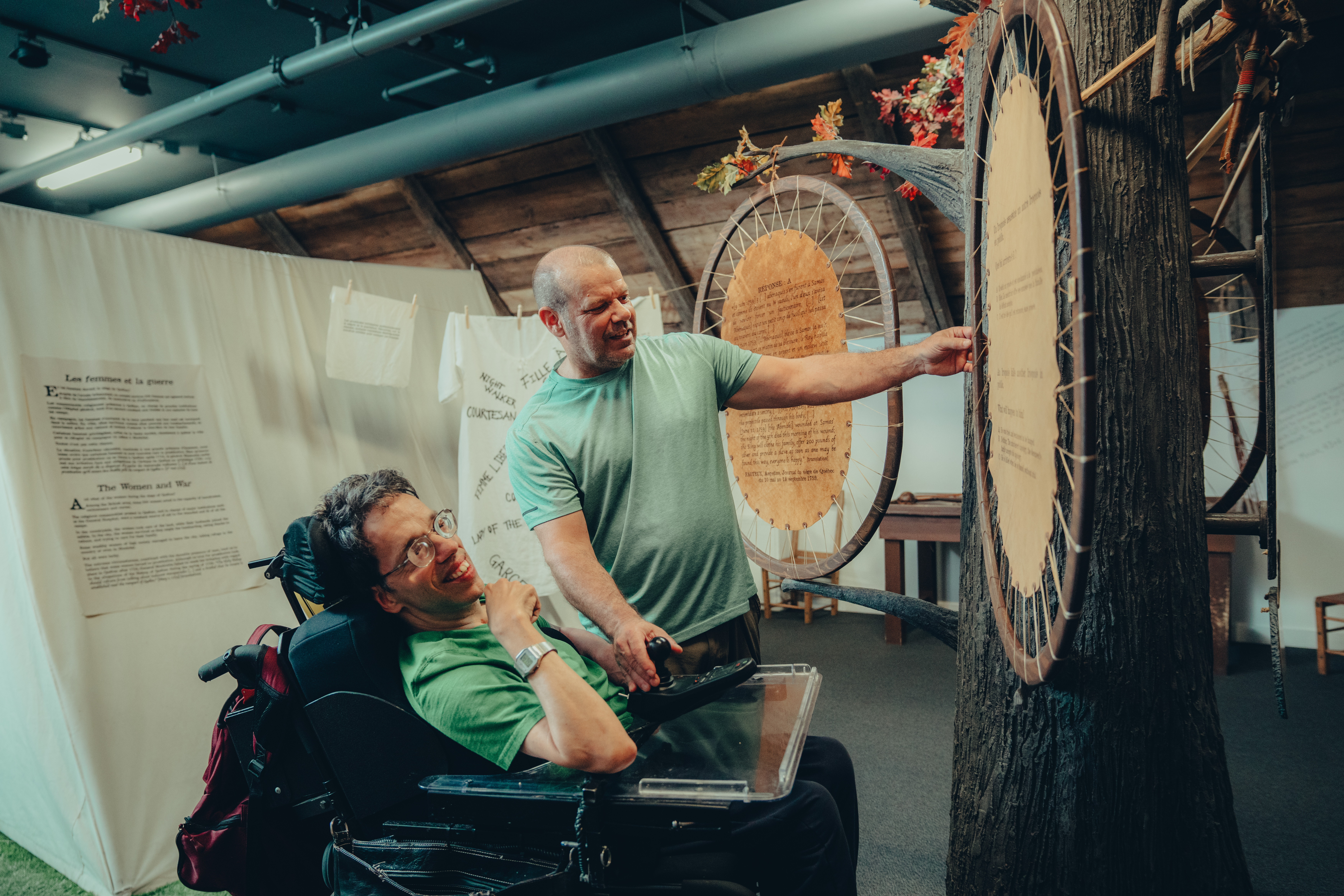 Un individu en fauteuil roulant et son accompagnateur contemple une pièce de l'exposition suspendu. An man in a wheelchair and their companion are admiring a suspended exhibition piece.