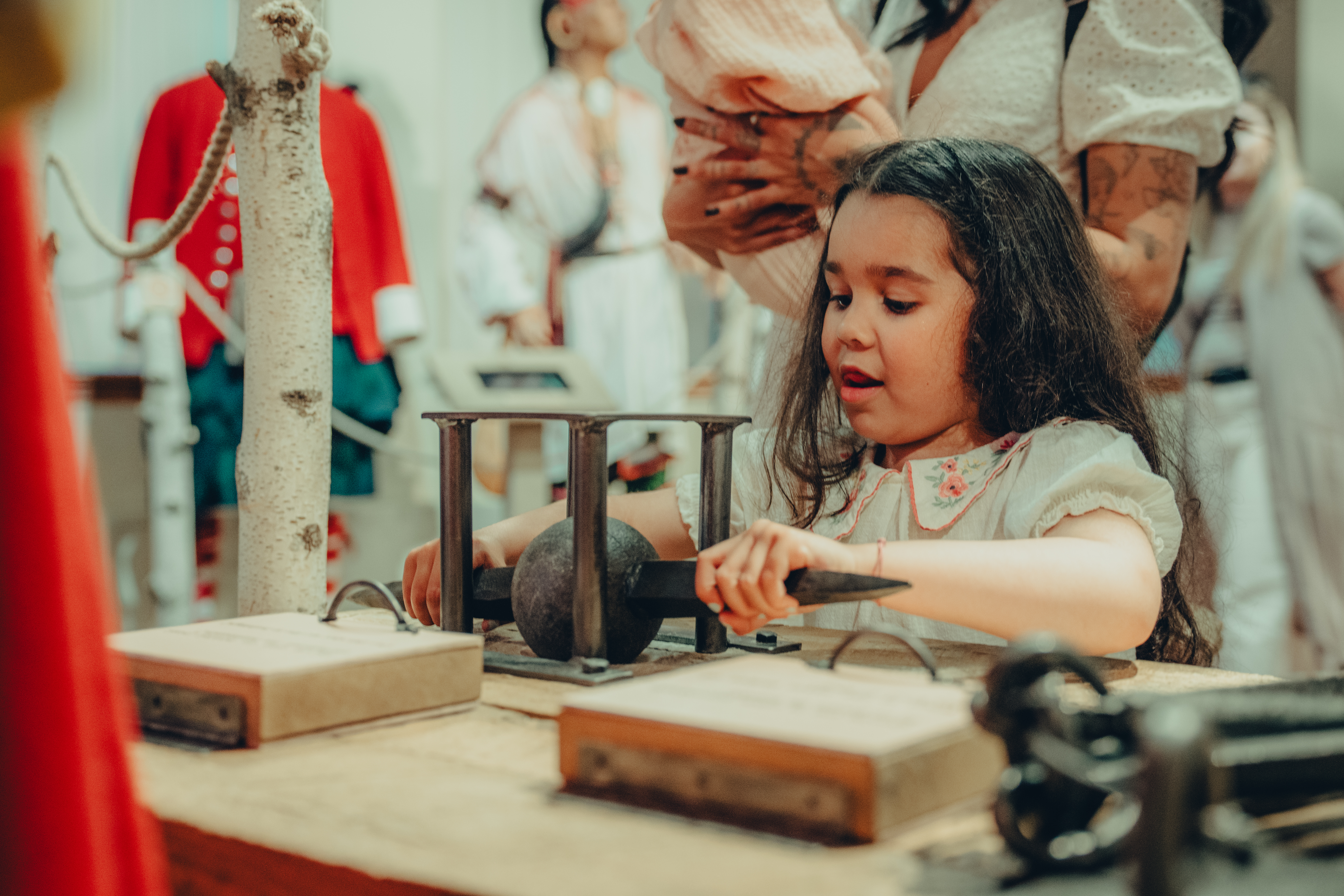 Fillette aux cheveux bruns, portant une robe beige, elle joue avec des instruments en bois déposés sur un présentoir. A young girl with brown hair, wearing a beige dress, playing with wooden instruments displayed on a stand.