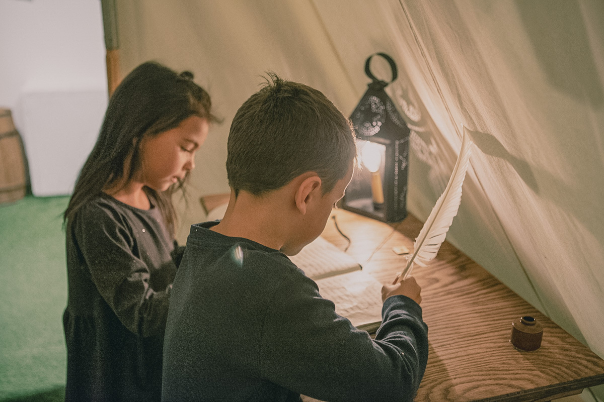 Un garçon et une fillette sous une tente en train de lire et d'écrire à l'aide d'une plume, une lanterne est placée sur la table de bois pour éclairer sous la tente. A boy and a girl under a tent reading and writing with a quill, with a lantern placed on the wooden table to provide light under the tent.