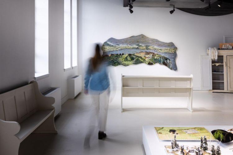 Photo d'une dame en train de marcher dans une exposition aux murs et au sol blanc, des oeuvres sont exposées sur le mur devant elle et au sol. Photo of a woman walking through an exhibition with white walls and floor, with artworks displayed on the wall in front of her and on the floor.